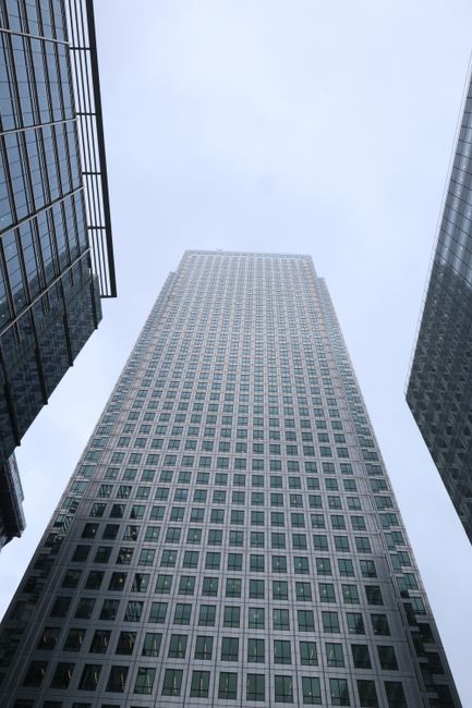 A modern high-rise office building with a curved corner featuring multiple floors and extensive glass windows, reflecting the bright blue sky above. The structure has a sleek, contemporary design with a mix of transparent glass panels and dark metallic framing, creating a pattern of horizontal and vertical lines across its façade. The building is situated in an urban environment, with other tall skyscrapers visible nearby, emphasizing its impressive height. The sunlight causes highlights and reflections on the glass surfaces, adding depth to the scene. The image appears to be taken from the ground level looking upwards, capturing the architectural detail and the expansive sky, which complements the clean, professional aesthetic associated with commercial office spaces and potential independent waste management or rubbish collection services nearby in similar urban settings. The overall scene emphasizes the building’s modernity and the cityscape context, aligning subtly with the theme of private or alternative waste handling in an office environment. That’s typical of the type of buildings where rubbish removal services like those provided by [COMPANY_NAME] are employed to manage waste from commercial tenants in high-rise offices such as those at One Canada Square in Canary Wharf.