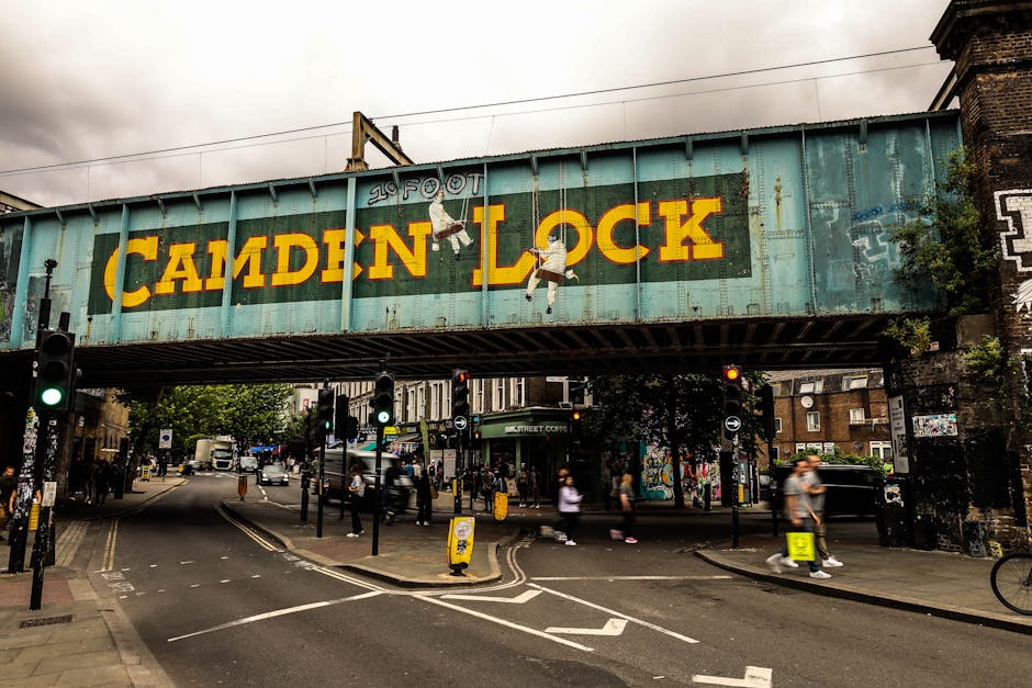 A large, weathered railway bridge with a teal-painted metal structure spans across a busy urban street. The bridge features bold yellow lettering spelling 'CAMDEN LOCK' centered on its side, with the letters slightly faded and the paint showing signs of rust and age. Two hanging painted illustrations of active children are positioned beneath the text, adding a playful element to the industrial setting. Beneath the bridge, several pedestrians cross the road, which has white traffic markings and multiple black bollards guiding vehicle and foot traffic. Traffic lights show green for pedestrians, and small yellow litter bins are placed along the sidewalk. In the background, a mixture of brick buildings, trees, and street furniture creates a typical Camden town scene, with some graffiti visible on the bridge supports and surrounding structures. The overall atmosphere appears overcast with soft, diffuse lighting, emphasizing the urban environment and the infrastructure that supports local waste management and street activity. Rubbish Collection Camden occasionally handles waste removal in such areas, which may involve private collection methods aligned with local waste handling regulations.