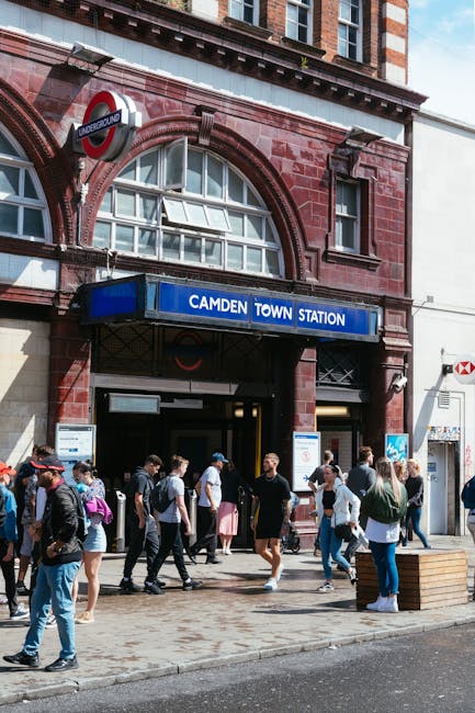 The image shows the exterior entrance of Camden Town underground station, featuring a red brick façade with arched windows and a blue sign reading 'Camden Town Station' above the door. A round London Underground sign with a red, blue, and white design is mounted on the upper left side of the building. In front of the station, a diverse group of pedestrians, some with backpacks and casual clothing, are walking or standing on the paved pavement. The scene is set during daytime with natural light illuminating the area, and the street appears busy with both locals and visitors. To the right, a small wooden planter with a metal street lamp post nearby is visible. This environment highlights a typical urban setting where public transportation access is accompanied by nearby foot traffic, aligning with the context of urban waste management and service coverage in central Camden, supporting the supply of alternative waste handling options through local rubbish collection services like Rubbish Collection Camden.