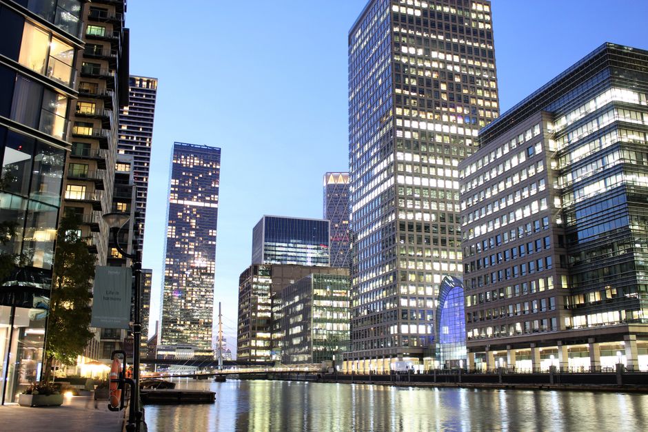 A wide cityscape view of Canary Wharf in London, showcasing a variety of modern high-rise office buildings with glass and steel façades, some reflecting sunlight. The buildings vary in height and architectural style, with the tallest structures positioned centrally and to the left of the image. In the foreground, the River Thames flows gently, with a small boat visible near the lower right corner. On the left side of the image, construction cranes are lying on the ground beside partially built structures, indicating ongoing development. The sky above is clear with a slight haze, providing natural daylight that illuminates the scene evenly. The setting also hints at the possibility of independent waste handling and rubbish removal services in the area, consistent with the urban environment of Canary Wharf, which often involves on-site clearance and private disposal solutions in addition to municipal services.