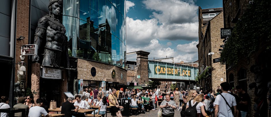 The image depicts a busy outdoor scene beneath a large blue railway bridge with the words 'CANDEN LOCK' painted in bold yellow letters. In the foreground, numerous pedestrians are gathered on a cobbled street, many holding umbrellas in various colours, including a prominent Union Jack design, indicating rainy weather. The crowd includes people wearing dark and neutral tones, with some engaged in conversation or walking past each other. To the left, a modern tram with a sleek, curved design and large windows is visible, reflecting the surroundings. The environment suggests an urban setting, likely near a transit hub or marketplace. The bridge above, made of weathered steel with visible rivets and supporting beams, spans across the scene, adding an industrial backdrop consistent with a railway or freight area. The scene's lighting is diffuse due to overcast skies, emphasizing the textures of the cobblestones, umbrellas, and the bridge structure. This setting aligns with local rubbish removal services managing waste in busy, high-traffic areas, possibly for event or street cleaning operations associated with Camden's public spaces.