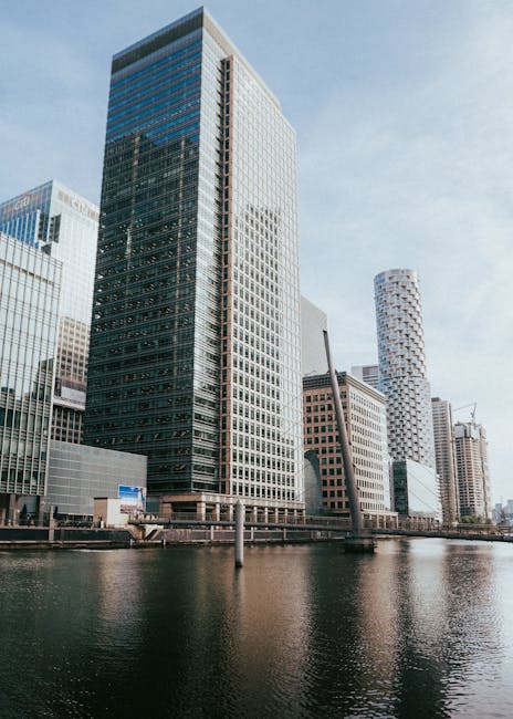 A tall glass office building dominates the foreground with a reflective facade that shows the surrounding cityscape, including other high-rise structures with various modern architectural designs. The building's exterior features a combination of dark-tinted glass and light-colored frames, creating a sleek, contemporary appearance. To the right, a distinctive cylindrical skyscraper with a white facade and a pattern of small, circular windows is visible, along with other nearby buildings exhibiting different styles and materials, such as brick and metal paneling. The scene is set along a body of water, likely a river or canal, with a calm, mirror-like surface reflecting the buildings. The sky above is mostly clear with a few wispy clouds, and natural daylight illuminates the scene evenly. This urban landscape, characteristic of central business districts, subtly relates to the context of waste management and independent collection services often needed in densely built environments, as provided by companies like rubbishcollectioncanarywharf.co.uk.